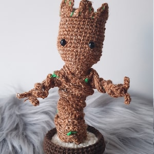 Peut inclure: Groot crocheté de Guardians of the Galaxy, debout dans un pot brun. Le personnage est brun avec des feuilles vertes et des yeux noirs.