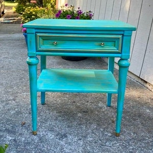 May include: A teal blue end table with a single drawer and a lower shelf. The table has turned legs and gold accents.