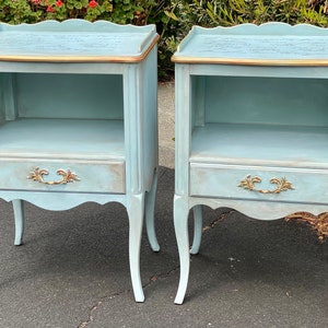 May include: A pair of light blue bedside tables with gold accents and a drawer. Each table has a shelf and a scalloped top.