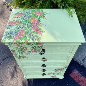 Puede incluir: Una c&oacute;moda verde claro con cuatro cajones y detalles florales. El dise&ntilde;o floral est&aacute; pintado en la parte superior y los lados de la c&oacute;moda.