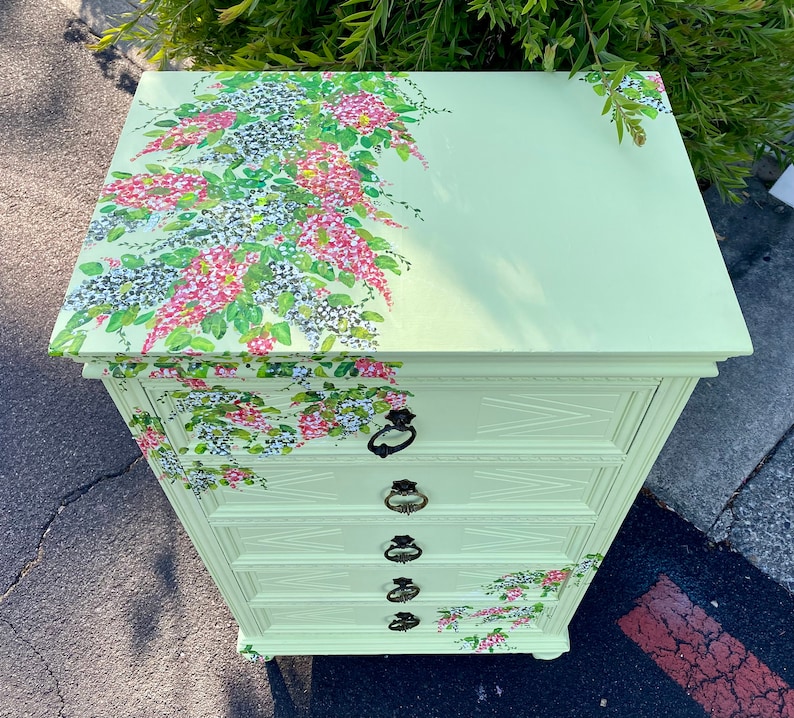 Puede incluir: Una c&oacute;moda verde claro con cuatro cajones y detalles florales. El dise&ntilde;o floral est&aacute; pintado en la parte superior y los lados de la c&oacute;moda.