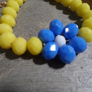 May include: A necklace featuring faceted yellow beads and a cluster of blue and white beads. The necklace has a gold-colored chain and clasp. The beads are arranged on a gray wooden surface.