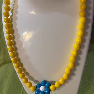 May include: A necklace featuring a continuous strand of round, bright yellow beads. The centre of the necklace is adorned with a single flower-shaped charm composed of six glossy blue petals surrounding a small, round, white centre bead.
