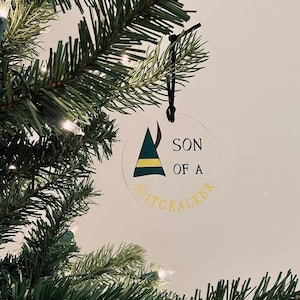 May include: A clear round Christmas bauble with a green and yellow elf hat and the text "SON OF A NUTCRACKER" in yellow lettering.