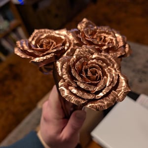 May include: A bouquet of copper roses with detailed petals. The metallic flowers have a warm, reddish-brown hue and are held together at the stems. The roses are a unique decorative item.