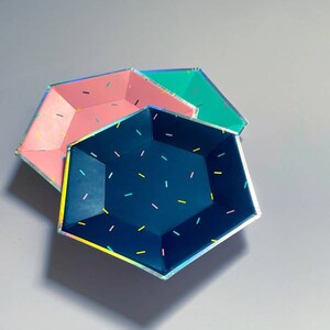 May include: Three hexagonal paper plates with a holographic finish. The plates are layered on top of each other. The top plate is dark blue with white and yellow sprinkles. The middle plate is pink with white sprinkles. The bottom plate is teal with white sprinkles.