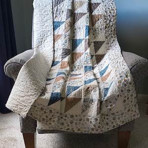 May include: A quilted blanket featuring a patchwork design of triangles in shades of blue, brown, and cream. The fabric includes patterns of small floral motifs and acorns. The quilt has a cream-colored border with a repeating acorn print.