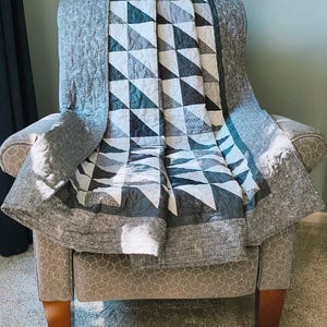 May include: A quilt featuring a geometric pattern of triangles in shades of charcoal gray, light gray, and white. The central design is framed by a solid gray border with a textured appearance, and the entire piece has visible machine quilting.
