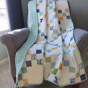 May include: A white quilt with a patchwork design featuring squares of blue, green, yellow, and white fabric. The quilt is folded and draped over a gray armchair.