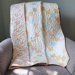 May include: A white and cream quilt with a floral pattern on one side and a patchwork pattern on the other side. The quilt is folded and sitting on a gray armchair.