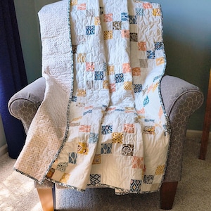 May include: A handmade quilt draped over a gray armchair. The quilt features a patchwork design with squares in shades of white, blue, orange, and yellow. The edges are finished with a patterned border. The chair has wooden legs.