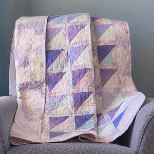 May include: A pastel-colored quilt draped over a gray armchair. The quilt features a pattern of triangles in shades of purple, pink, and yellow. The edges of the quilt have a floral design. The quilt is made of soft fabric.