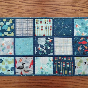 May include: A rectangular quilted placemat with a patchwork design. The squares feature various patterns, including trees, oars, floral prints, and birds, in shades of blue, teal, and white. The placemat is bordered with a dark blue fabric.