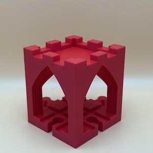 May include: A red, cube-shaped toy castle with crenelated battlements. The toy has arched openings on each side and a detailed, geometric design. The toy is made of a solid material and sits on a white surface.