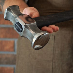 May include: A silver metal hammer with a dark handle. The hammer head has a flat striking face and a rounded peen. The handle is textured for grip. The hammer is held in a hand.