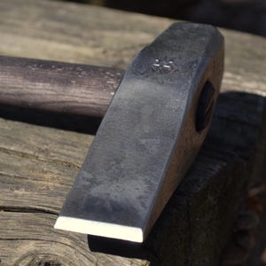 Peut inclure: Un marteau en métal noir avec un manche en bois. Le marteau a une tête plate et un manche légèrement incurvé. Le marteau est posé sur une surface en bois.