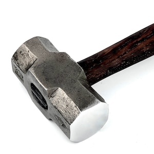 May include: A heavy sledgehammer featuring a dark, textured metal head and a polished striking face. It has a dark, stained wooden handle. This tool is designed for demolition and heavy construction tasks.