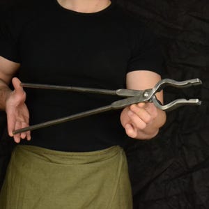 Può includere: Un fabbro tiene in mano un paio di tenaglie lunghe in metallo con una ganascia curva. Le tenaglie sono in acciaio e hanno un aspetto rustico, forgiato a mano.
