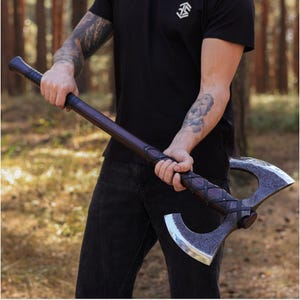 May include: A handcrafted Viking axe with a dark wooden handle and a double-bladed, silver-colored head. The handle is wrapped with dark leather and secured with metal studs. The axe is held against a blurred forest background.