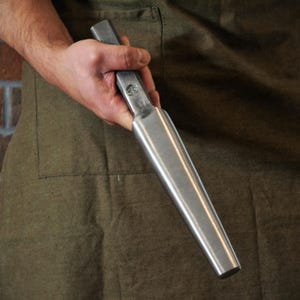 May include: A metal tool with a rectangular handle and a tapered, cylindrical body. The tool is held against a green apron. The tool appears to be made of steel and has a smooth, reflective surface.