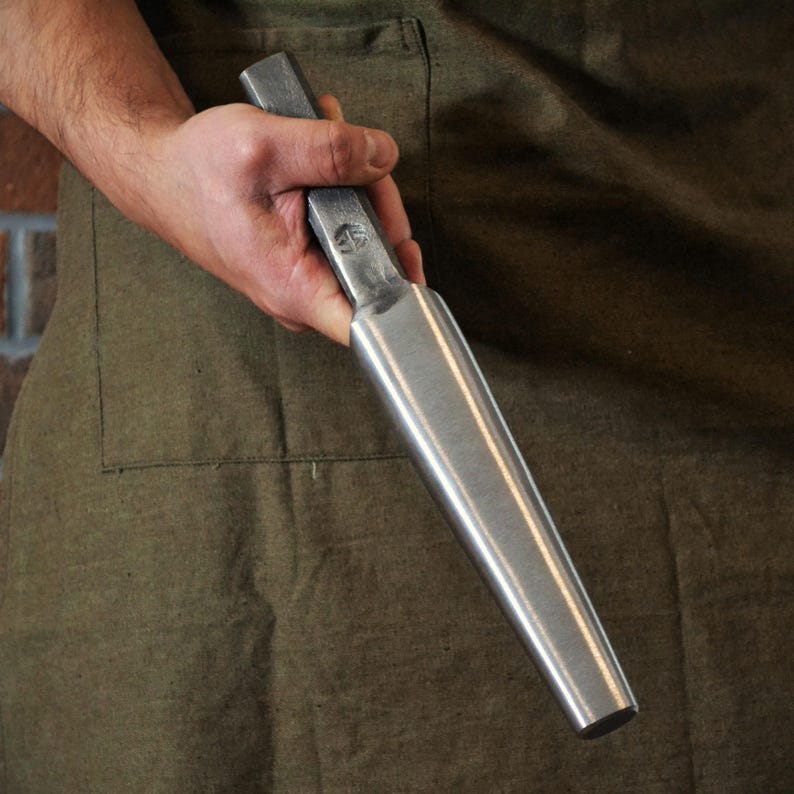 May include: A metal tool with a rectangular handle and a tapered, cylindrical body. The tool is held against a green apron. The tool appears to be made of steel and has a smooth, reflective surface.