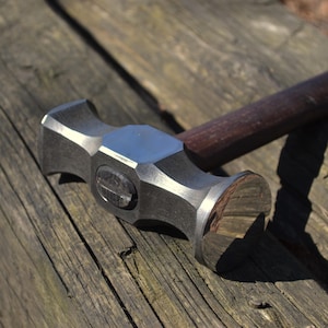 Pode incluir: Um martelo de metal com um cabo de madeira escura. A cabeça do martelo é uma combinação de metal polido e texturizado, com uma face de impacto plana. O cabo é de cor castanho escuro e o martelo repousa sobre uma superfície de madeira envelhecida.