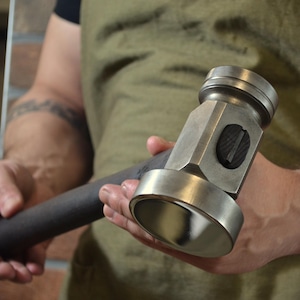 May include: A steel hammer with a dark handle. The hammer head has a flat striking face and a rounded face. The hammer is held by a person wearing a green apron.