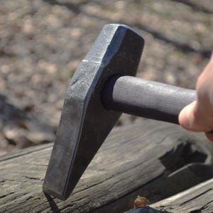 May include: A close-up of a black metal hammer with a wooden handle. The hammer is resting on a piece of wood.