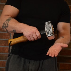 May include: A silver and black hammer with a wooden handle. The hammerhead features an intricate Celtic knot design. The handle is made of light brown wood and is attached to the hammerhead with a black leather grip.