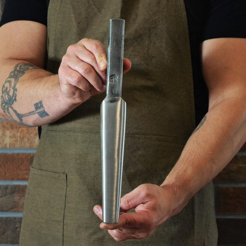 May include: A metal tool with a rectangular top and a tapered, cylindrical body. The tool is held by a person wearing a brown apron. The tool has a small circular marking on the top.