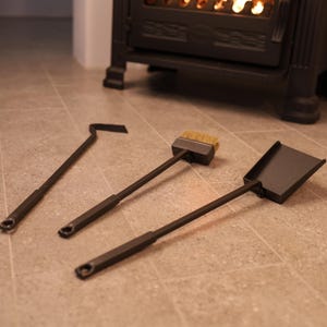 May include: A set of three fireplace tools: a poker, a brush, and a shovel. The tools are made of black metal and have wooden handles. The tools are lying on a light brown tiled floor.