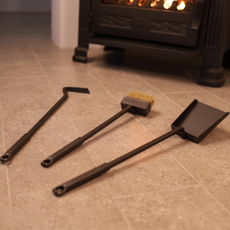 May include: A set of three fireplace tools: a poker, a brush, and a shovel. The tools are made of black metal and have wooden handles. The tools are lying on a light brown tiled floor.