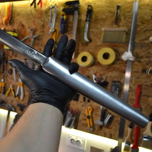 Puede incluir: Una varilla de metal plateado con un extremo redondeado y un extremo plano cuadrado. La varilla mide aproximadamente 45 cm de largo y tiene un ligero estrechamiento.