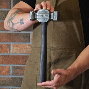 May include: A blacksmith's hammer with a dark wooden handle and a silver-colored head. The hammer's head has a flat striking surface and a rounded peen. The handle is long and cylindrical. The hammer is held in front of a brick wall.