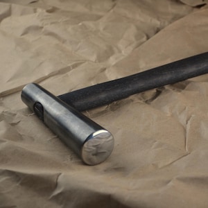 Könnte beinhalten: Ein Metallhammer mit zylindrischem Kopf und dunklem Holzgriff. Der Hammerkopf ist silberfarben und der Griff ist schwarz. Der Hammer liegt auf einer braunen Papieroberfläche.