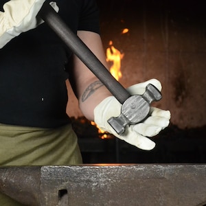 Könnte beinhalten: Ein Schmiedehammer mit einem dunklen, texturierten Metallgriff und einem schweren Kopf aus geschmiedetem Stahl, der ein markantes Kreuzfinne-Design aufweist. Das Werkzeug hat eine rohe, industrielle Oberfläche mit sichtbaren Hammerschlägen.