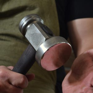 May include: A metal hammer with a dark wooden handle and a round, pink-colored striking face. The hammer's head is silver and has a square shape. The tool is held in a person's hands.