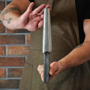 May include: A metal tool with a tapered, cylindrical head and a rectangular handle. The tool is held against a brick wall. The tool has a logo stamped on the handle. The tool is silver in color.