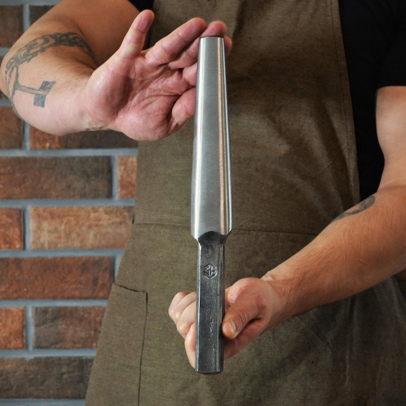 May include: A metal tool with a tapered, cylindrical head and a rectangular handle. The tool is held against a brick wall. The tool has a logo stamped on the handle. The tool is silver in color.