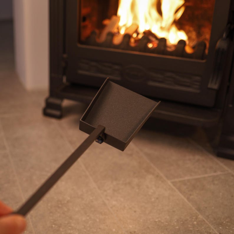 May include: A black metal fireplace shovel with a long handle. The shovel is being used to remove ashes from a fireplace.