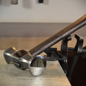 Pode incluir: Um martelo de metal prateado com cabo de madeira. O martelo tem duas cabeças de formas diferentes, uma redonda e uma quadrada. O martelo está apoiado numa superfície cinzenta.