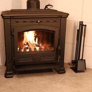 May include: A black cast iron wood-burning stove with a fire burning inside. The stove has a decorative design on the front and a small metal dragon on top. A set of black fireplace tools is leaning against the wall to the right of the stove.