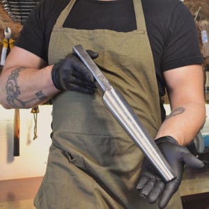 May include: A long, silver metal tool with a square head and a tapered, pointed end. The tool is held in gloved hands.