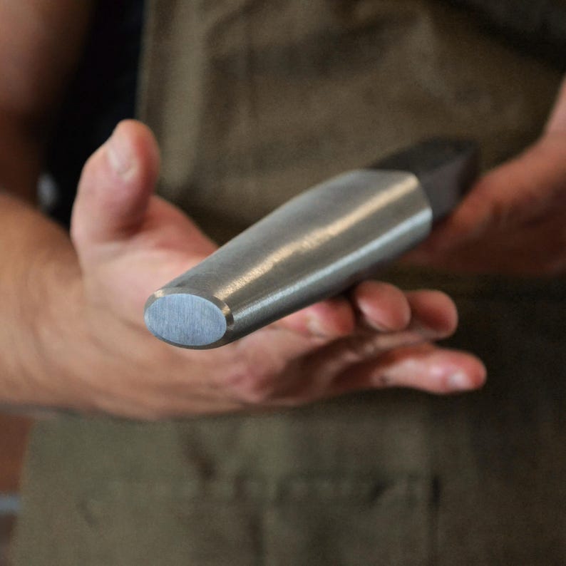 May include: A close-up of a metal tool with a rounded, oval end and a cylindrical body. The tool has a dark, rectangular section at the opposite end. The tool is held in a person's hands.