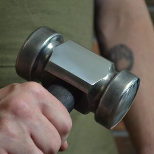 Könnte beinhalten: Ein Metallhammer mit einem dunklen Holzgriff. Der Hammerkopf ist silberfarben mit einer flachen Schlagfläche. Der Hammer wird in der Hand einer Person gehalten.