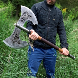 Puede incluir: Un hacha grande de doble hoja con un mango de madera y diseños grabados intrincados en las hojas de acero. El mango está envuelto en cuero marrón. El hacha se sostiene frente a un fondo de follaje verde.