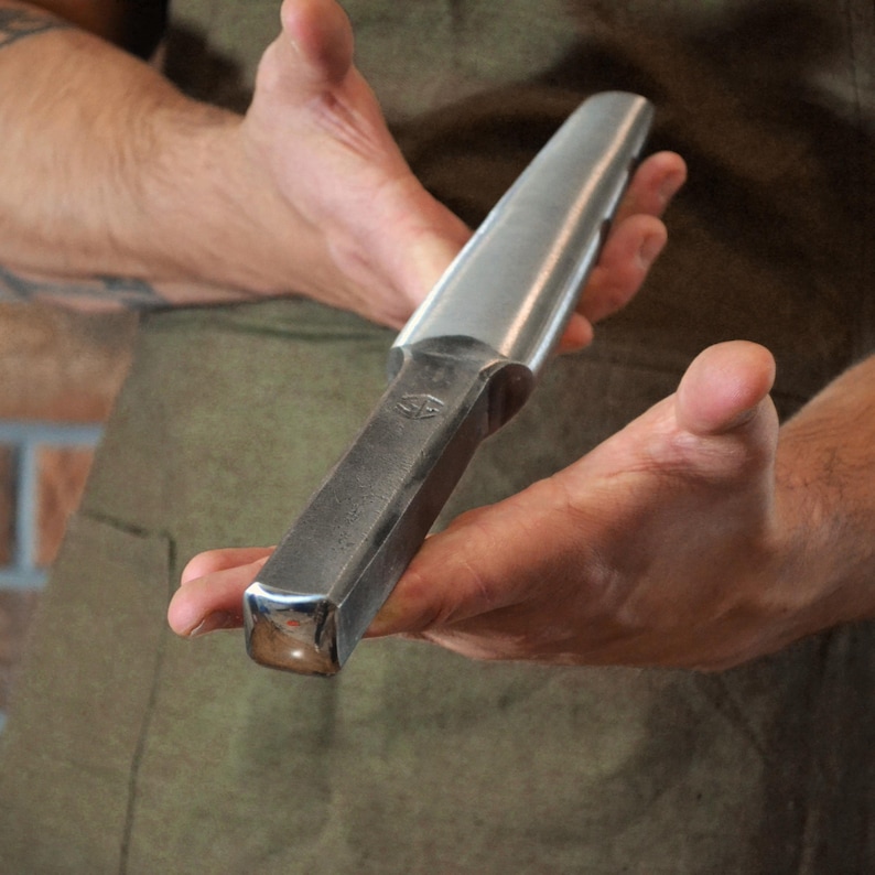 May include: A metal chisel with a rectangular handle and a curved, polished blade. The tool is held in two hands, showcasing its design and craftsmanship. The chisel appears to be a woodworking or metalworking tool.