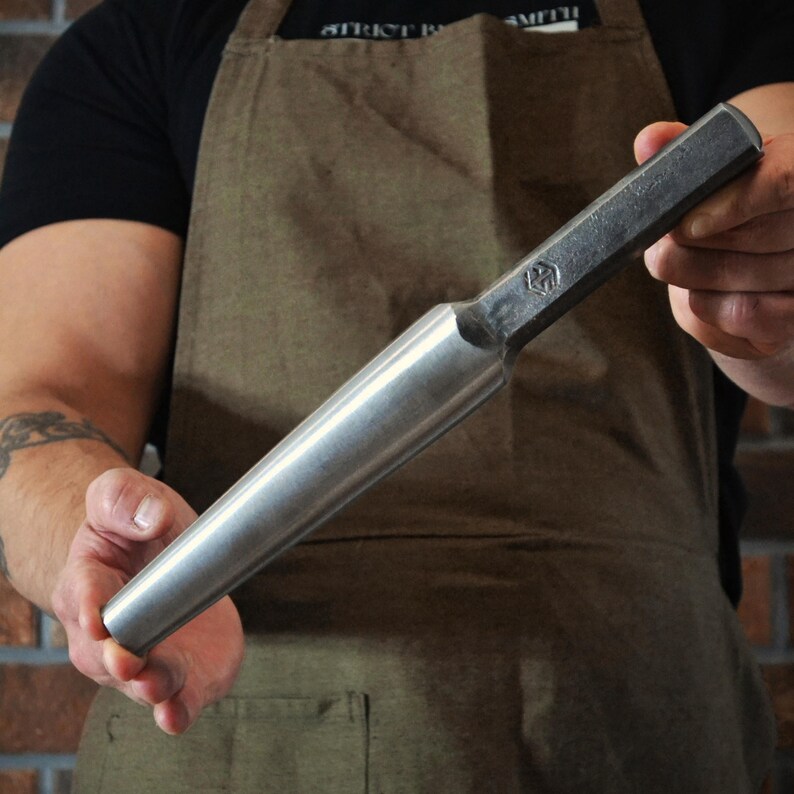 May include: A metal tool with a cylindrical handle and a hexagonal head. The tool has a silver finish on the handle and a darker, textured finish on the head. The tool is held by a person wearing a brown apron.