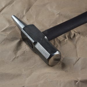 Peut inclure: Un marteau en acier avec un manche en bois noir. La tête du marteau est gris foncé avec une face de frappe plate et une panne conique. Le marteau est posé sur une surface en papier brun froissé.
