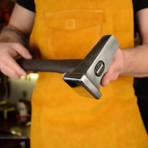 May include: A person holding a large, silver hammer with a wooden handle. The hammer has a square head and a flat striking surface.